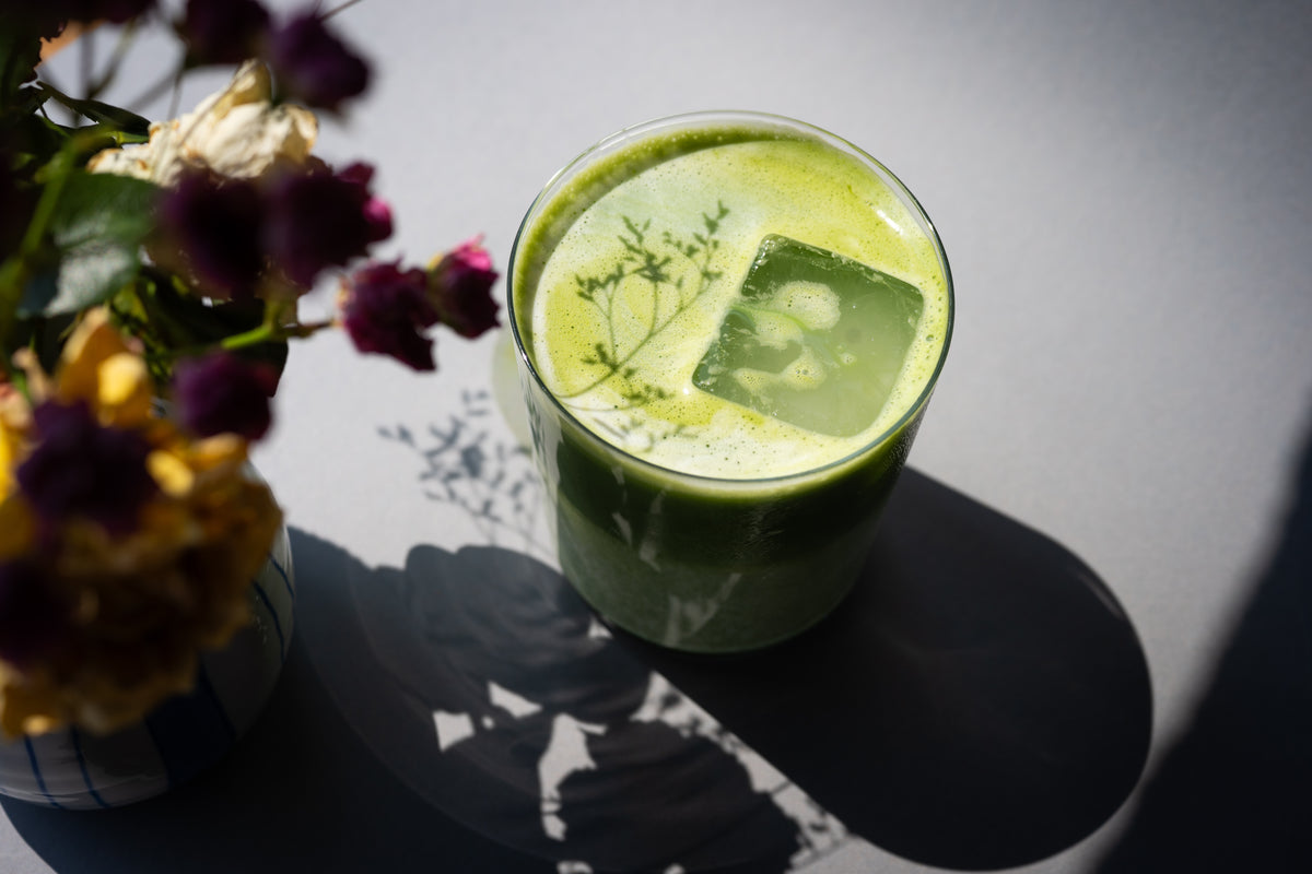 Iced Matcha Latte mit cremigem Schaum und Eiswürfel, serviert in einem Glas auf grauem Tisch mit Schatten von Blumen
