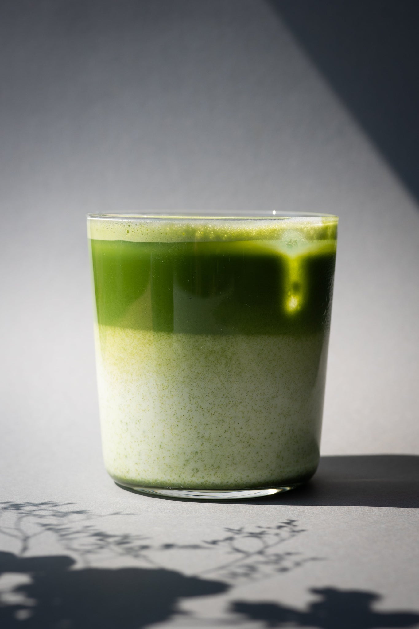Schichtiger Iced Matcha Latte in einem Glas, unten cremige Milch, oben leuchtend grüner Matcha, im Sonnenlicht mit Schatten von Pflanzen im Vordergrund