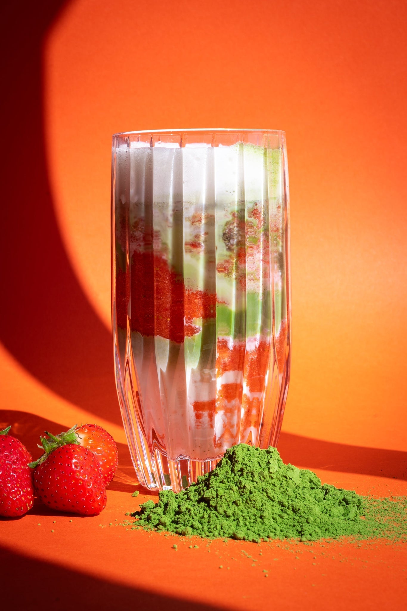 Strawberry Iced Matcha Latte im hohen Glas, geschichtet mit Milch, pürierten Erdbeeren und grünem Matcha, daneben frische Erdbeeren und Matcha-Pulver vor orangem Hintergrund