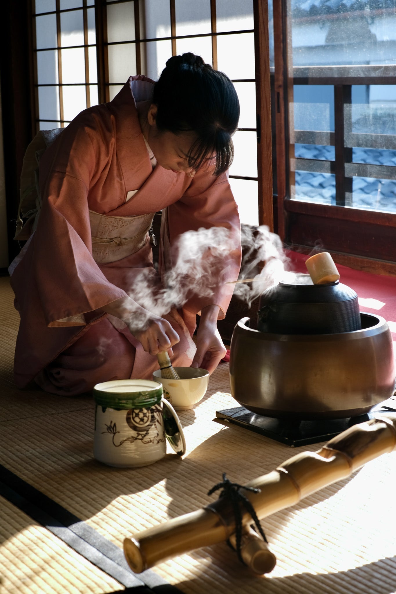 Japanische Frau im Kimono bereitet traditionell Matcha-Tee im Rahmen einer Teezeremonie zu, kniend auf Tatami-Matten mit dampfendem Wassergefäß und Chasen (Bambusbesen) in der Hand.