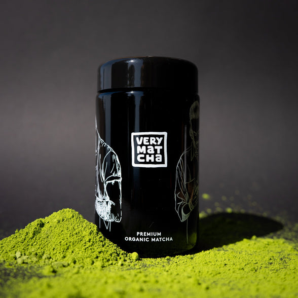 Bio Matcha Pulver mit schwarzem Very Matcha Glas im Vordergrund auf dunklem Hintergrund.