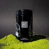 Bio Matcha Pulver mit schwarzem Very Matcha Glas im Vordergrund auf dunklem Hintergrund.