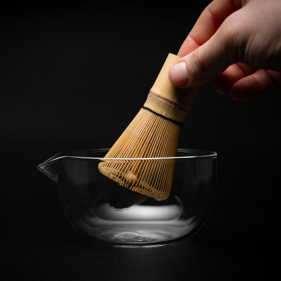 Matcha Schale aus Glas mit Ausgießer, in die ein traditioneller Matcha-Besen aus Bambus (Chasen) gehalten wird, vor schwarzem Hintergrund.
