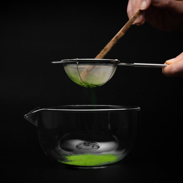 Zubereitung in der Matcha Schale aus Glas: Ein Metallsieb wird über die Schale gehalten, während Matcha-Pulver mit einem Bambuslöffel hineingesiebt wird.