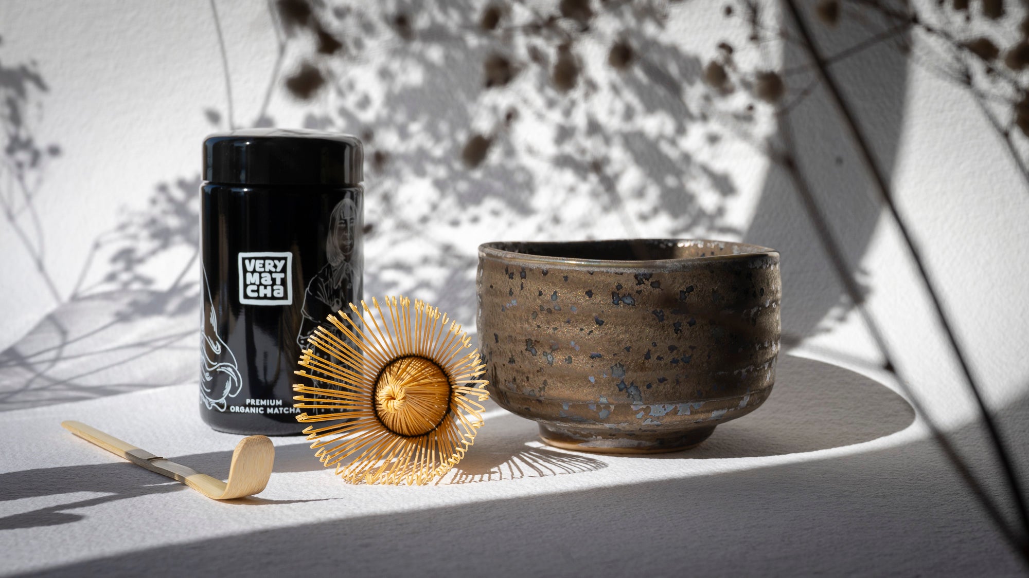 Die Matcha Schale Hoshiakari zusammen mit dem Ceremony Bio Matcha, Bambuslöffel und einem Matcha-Besen vor weißem Hintergrund mit floralen Schatten.